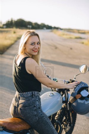 Photo for Biker girl sits on a motorcycle. Young beautiful blonde woman sitting on a motorcycle on the highway at sunset. - Royalty Free Image