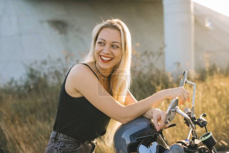 Téléchargez les photos : Une motard est assise sur une moto. Jeune belle femme blonde assise sur une moto sur l'autoroute au coucher du soleil. - en image libre de droit