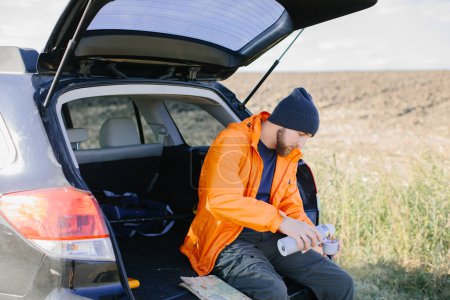 Foto de Un hombre descansa sentado en el maletero de un coche, bebiendo café de un termo. - Imagen libre de derechos