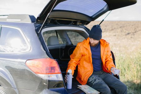 Photo for A man rests sitting in the trunk of a car, drinking coffee from a thermos. - Royalty Free Image
