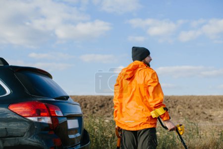 Photo for A man with a metal detector and a shovel in his hands stands near his car on the background of a field. Ready for a treasure hunt. - Royalty Free Image