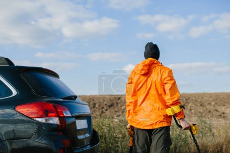 Foto de Un hombre con un detector de metales y una pala en sus manos está cerca de su coche en el fondo de un campo. Listo para una búsqueda del tesoro. - Imagen libre de derechos