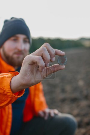 Un hombre tiene una moneda antigua en sus manos, encontrada en un campo con un detector de metales.