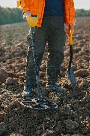 Una bobina de una pala detector de metales y las piernas de una persona que busca un tesoro sobre el fondo de un campo arado.