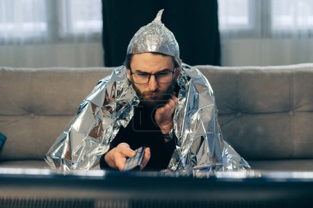 Foto de Un hombre en un sombrero de papel de aluminio mira la televisión. - Imagen libre de derechos