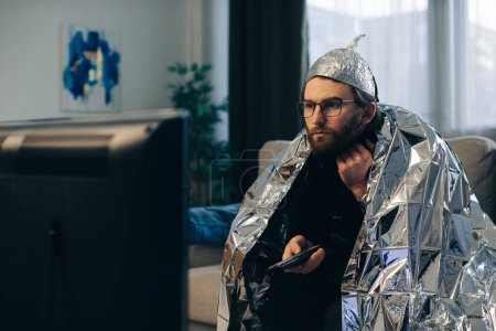 Photo for A young man wearing a tinfoil hat and wrapped in a tinfoil blanket is sitting on the couch in front of the TV with a remote control in his hands. - Royalty Free Image