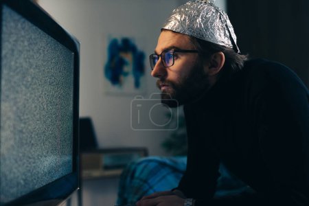 Photo for Conspiracy Theory on Screen: Man in Tin Foil Hat and Blanket Observing TV Interference - Royalty Free Image