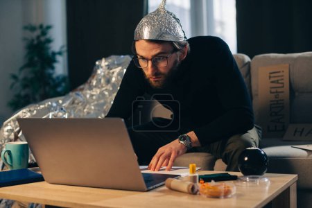 Théorie du complot. Un homme avec un chapeau en papier d'aluminium cherche des signes en regardant une vidéo sur un ordinateur portable.