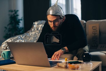 Foto de Teoría de la conspiración. Un hombre en un sombrero de papel de aluminio busca señales mientras ve un video en una computadora portátil. - Imagen libre de derechos