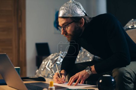 Foto de Teoría de la conspiración. Un hombre en un sombrero de papel de aluminio busca señales mientras ve un video en una computadora portátil. - Imagen libre de derechos