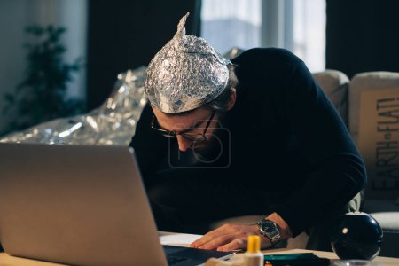 Théorie du complot. Un homme avec un chapeau en papier d'aluminium cherche des signes en regardant une vidéo sur un ordinateur portable.
