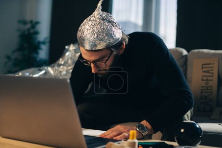 Téléchargez les photos : Théorie du complot. Un homme avec un chapeau en papier d'aluminium cherche des signes en regardant une vidéo sur un ordinateur portable. - en image libre de droit