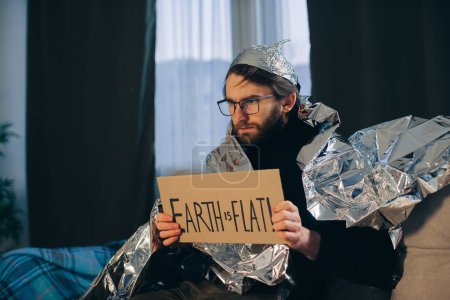 Foto de Teoría de la conspiración, un hombre envuelto en una manta de aluminio sostiene un letrero con una inscripción. - Imagen libre de derechos