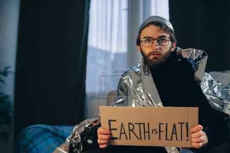 Flat Earth Advocate: Mann mit Blechfolienhut und Decke hält Schild "Die Erde ist flach"