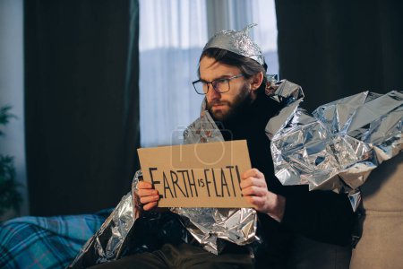 Photo for Flat Earth Advocate: Man in Tin Foil Hat and Blanket Holds Sign 'Earth is Flat - Royalty Free Image