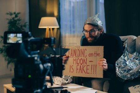 A conspiracy theorist shoots pseudoscientific videos on camera. A man in a tinfoil hat and a sign in his hands sits on a couch in front of the camera.