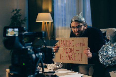 A conspiracy theorist shoots pseudoscientific videos on camera. A man in a tinfoil hat and a sign in his hands sits on a couch in front of the camera.