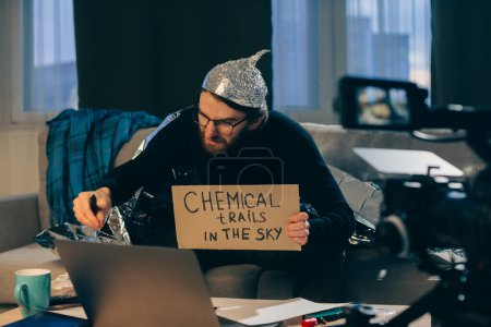 Un teórico de la conspiración dispara videos pseudocientíficos en cámara. Un hombre con un sombrero de papel de aluminio y un cartel en sus manos se sienta en un sofá frente a la cámara.