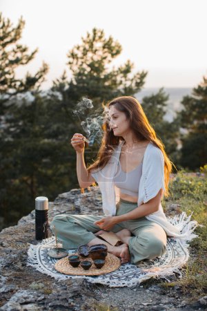 Foto de Una joven meditando con un palo santo fumando en sus manos mientras está sentada en una roca al atardecer. - Imagen libre de derechos
