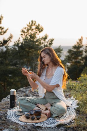 Photo for A young woman meditating with a smoking palo santo in her hands while sitting on a rock at sunset. - Royalty Free Image