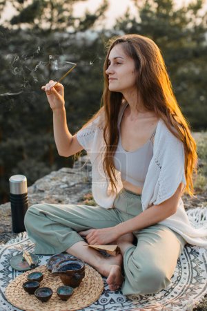 Foto de Una joven medita con una varilla de incienso en las manos mientras está sentada en una roca al atardecer. - Imagen libre de derechos