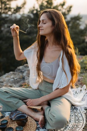 Foto de Una joven medita con una varilla de incienso en las manos mientras está sentada en una roca al atardecer. - Imagen libre de derechos