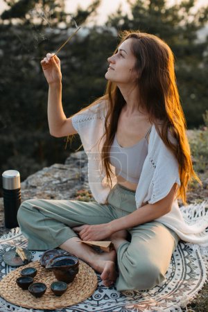 A young woman meditates with an incense stick in her hands while sitting on a rock at sunset.