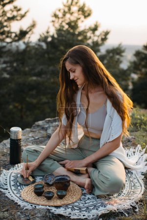 Téléchargez les photos : Une jeune femme médite avec un bâton d'encens dans les mains alors qu'elle est assise sur un rocher au coucher du soleil. - en image libre de droit