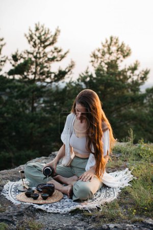 A young beautiful woman performs a Chinese tea ceremony in the mountains at sunset.