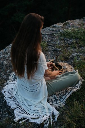 Foto de Mujer joven sentada en posición de loto con cuenco tibetano meditando en la montaña rocosa al atardecer. - Imagen libre de derechos