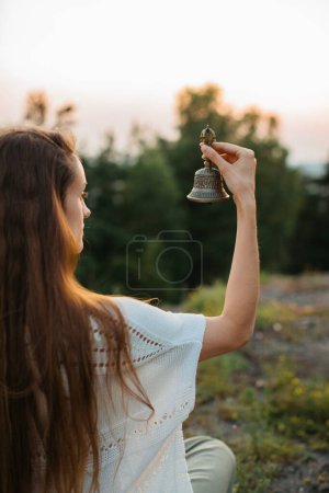 Téléchargez les photos : Une jeune femme méditant avec une cloche bouddhiste Gantha dans les mains, assise sur un rocher dans les rayons du soleil couchant. - en image libre de droit