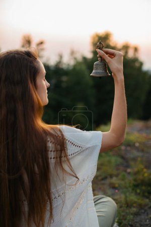 Photo for A young woman meditating with a Gantha Buddhist bell in her hands, sitting on a rock in the rays of the setting sun. - Royalty Free Image