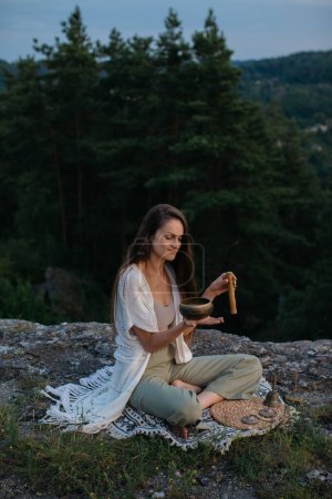 A young woman meditates with a tibetan singing bowl at sunset in the mountains.