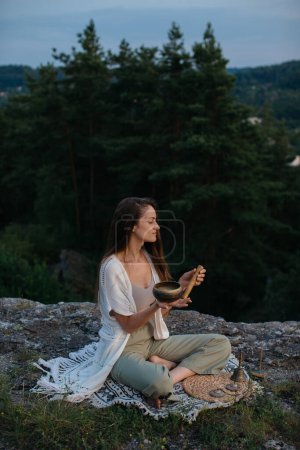 Une jeune femme médite avec un bol de chant tibétain au coucher du soleil dans les montagnes.