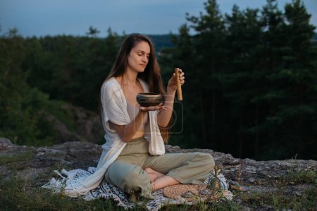 Photo for A young woman meditates with a tibetan singing bowl at sunset in the mountains. - Royalty Free Image