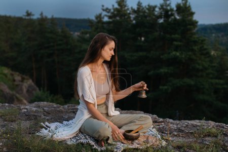 Foto de Una joven medita con una campana de Gantha mientras está sentada en una roca en los rayos del sol poniente. - Imagen libre de derechos