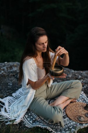 Foto de Joven morena meditando con un cuenco cantando al atardecer en las montañas. - Imagen libre de derechos