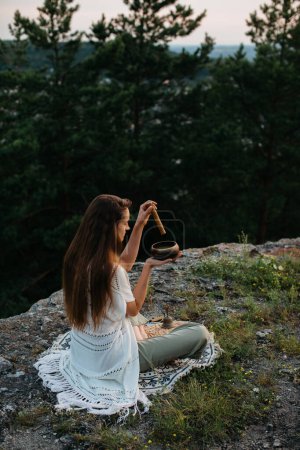 Foto de Una joven morena hermosa medita en las montañas en el fondo de un bosque de pinos en los rayos del sol poniente. - Imagen libre de derechos