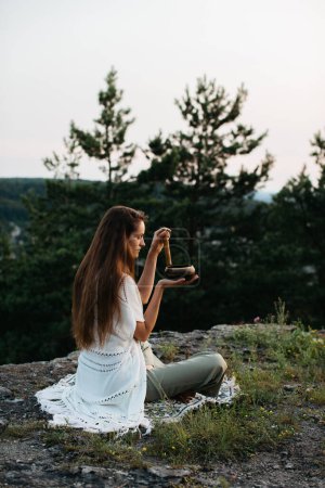 A young beautiful brunette woman meditates in the mountains on the background of a pine forest in the rays of the setting sun.
