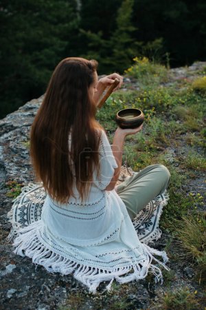 Foto de Una joven morena hermosa medita en las montañas en el fondo de un bosque de pinos en los rayos del sol poniente. - Imagen libre de derechos