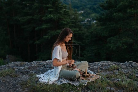 Photo for A young beautiful brunette woman meditates in the mountains on the background of a pine forest in the rays of the setting sun. - Royalty Free Image