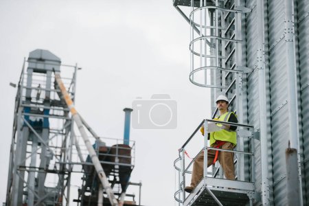 Foto de Un trabajador industrial masculino se para en un silo de grano con documentos en sus manos y hace una inspección visual. - Imagen libre de derechos