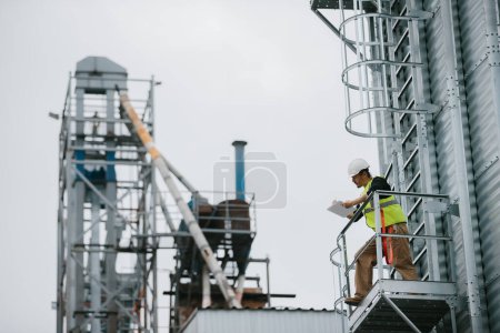 Photo for A male industrial worker stands on a grain silo with documents in his hands and makes a visual inspection. - Royalty Free Image