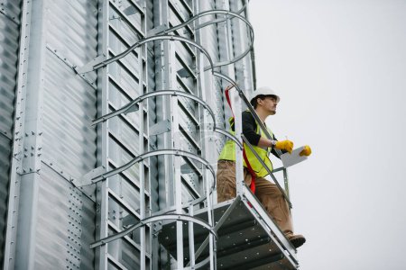 Foto de Un trabajador industrial masculino se para en un silo de grano con documentos en sus manos y hace una inspección visual. - Imagen libre de derechos