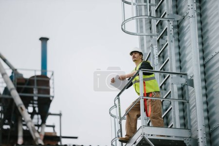 Téléchargez les photos : Un ouvrier industriel se tient debout sur un silo à grains avec des documents à la main et fait une inspection visuelle. - en image libre de droit