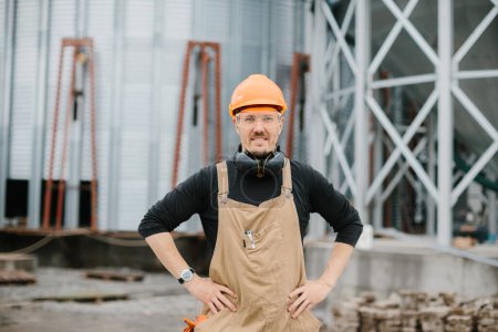 Photo for A builder in a protective helmet, glasses and overalls stands on the background of the construction of a grain silo. - Royalty Free Image