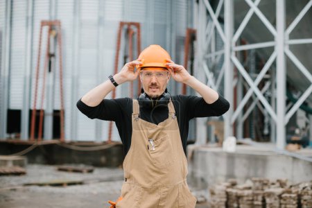 Foto de Un constructor en un casco de protección, gafas y monos se encuentra en el fondo de la construcción de un silo de grano. - Imagen libre de derechos