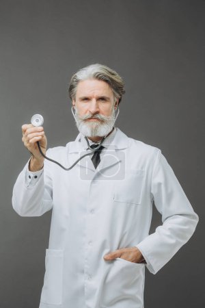 Photo for A gray-haired male doctor in a white medical coat with a phallendoscope in an outstretched hand on a gray background. - Royalty Free Image