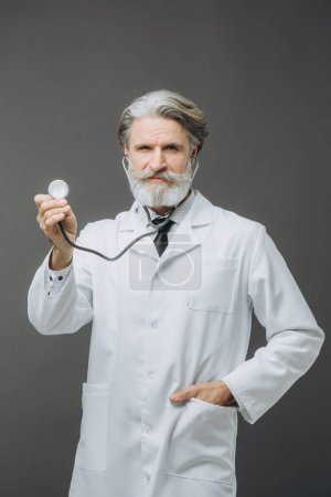 Téléchargez les photos : Un médecin aux cheveux gris dans un manteau médical blanc avec un phallendoscope dans une main tendue sur un fond gris. - en image libre de droit