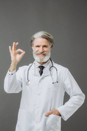 Photo for Gray-haired middle-aged man in doctor's coat with stethoscope, showing ok gesture, isolated on gray background. - Royalty Free Image